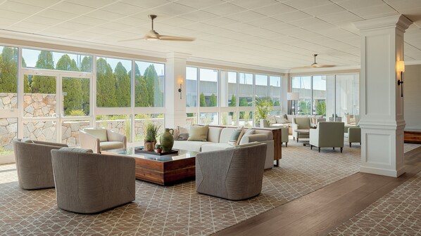 Un salón amplio con ventanas grandes, ventilador de techo y una variedad de opciones de asiento, incluyendo sofás y sillones.