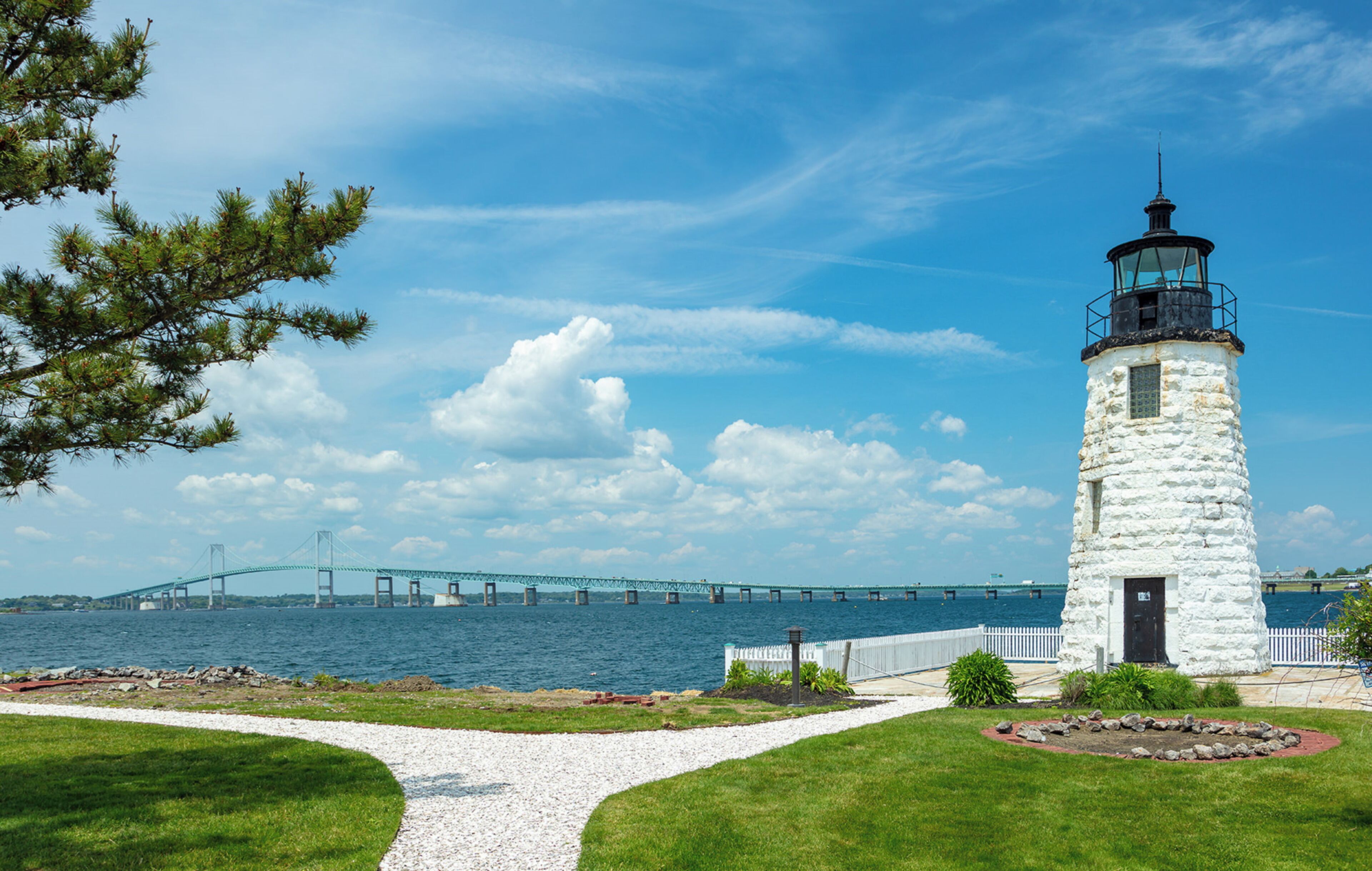 A lighthouse with a white exterior and black top, situated by a waterfront with a bridge in the background.