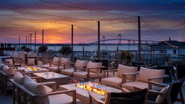 A deck with outdoor seating, a fire feature, and a view of a bridge and water at sunset. - Newport Harbor Island Resort (Newport)