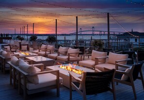 Una terraza con mobiliario exterior, un hogar para fuego y vistas a un puente y al agua al atardecer.