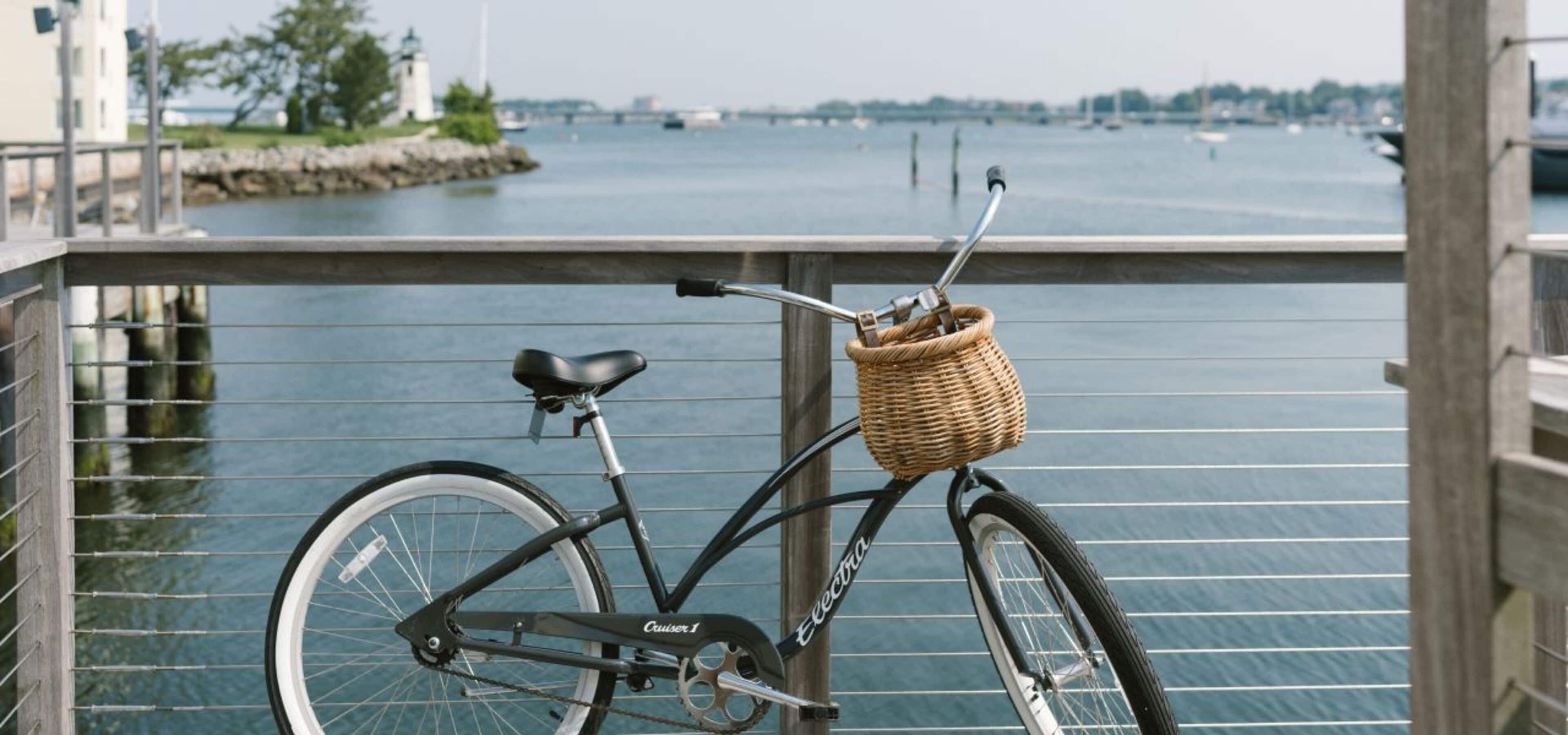 A bicycle with a wicker basket attached to the front, parked on a dock with a view of a body of water and a lighthouse in the background.