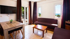 A room with a wooden table, a sofa, a TV mounted on the wall, and a view of the sea through a window. - Romanos Beach Villas (Pylos-Nestoras)