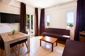 A room with a wooden table, a sofa, a television, and a view of the sea through a window. - Romanos Beach Villas (Pylos-Nestoras)