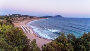 Uma praia com fileiras de guarda-sóis e cadeiras de praia, um litoral rochoso e uma montanha ao longe.