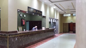 Un mostrador de recepción con un letrero que dice "RECEPTION" en inglés y otro idioma. Hay relojes en la pared, una pantalla digital y un cartel de no fumar.