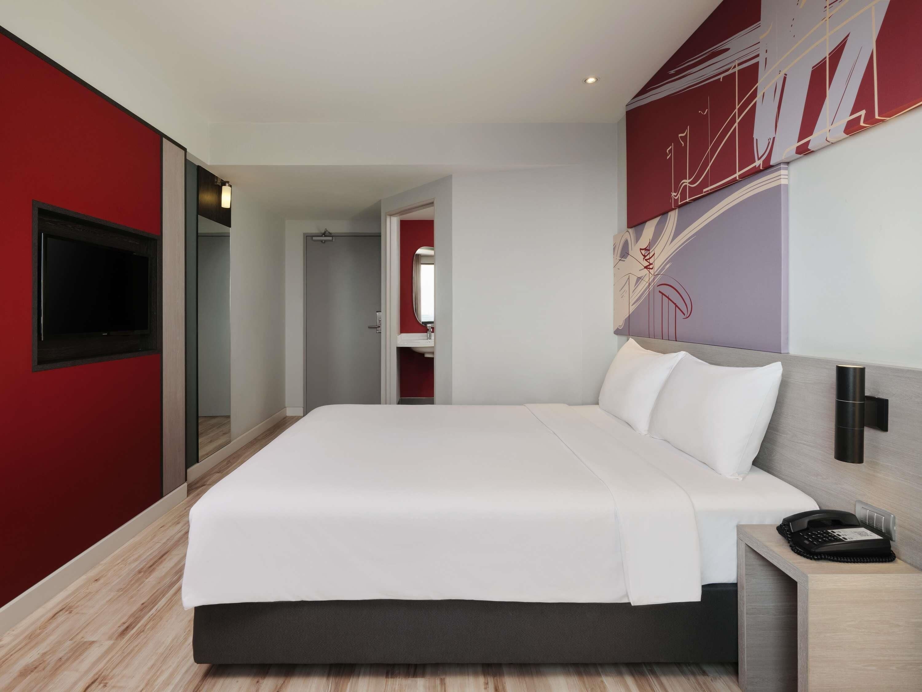 A modern hotel room with a large bed, a flat-screen TV, a bedside table, and a red accent wall.