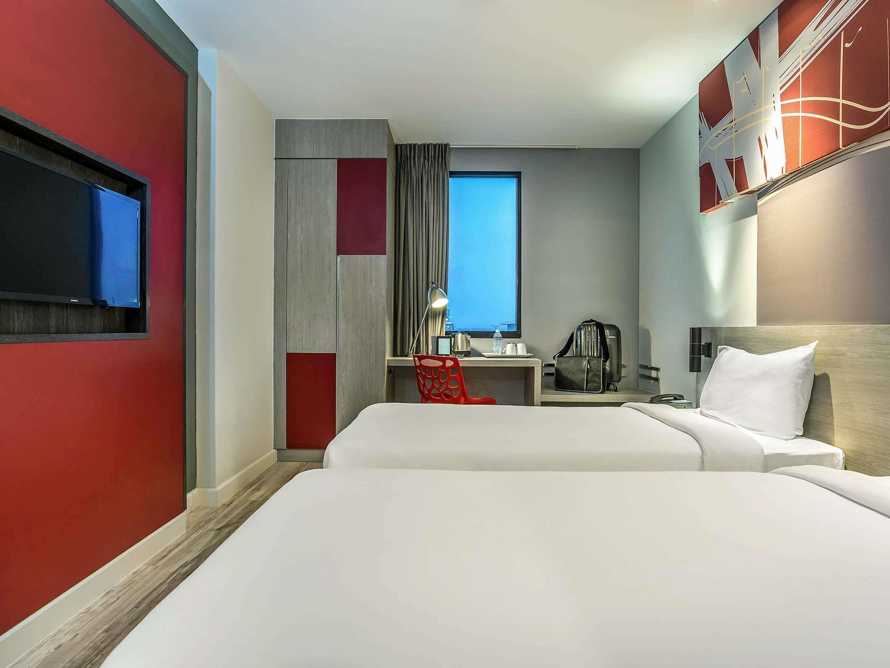 A hotel room with two beds, a desk with a chair, a TV, a window with curtains, and a city view.