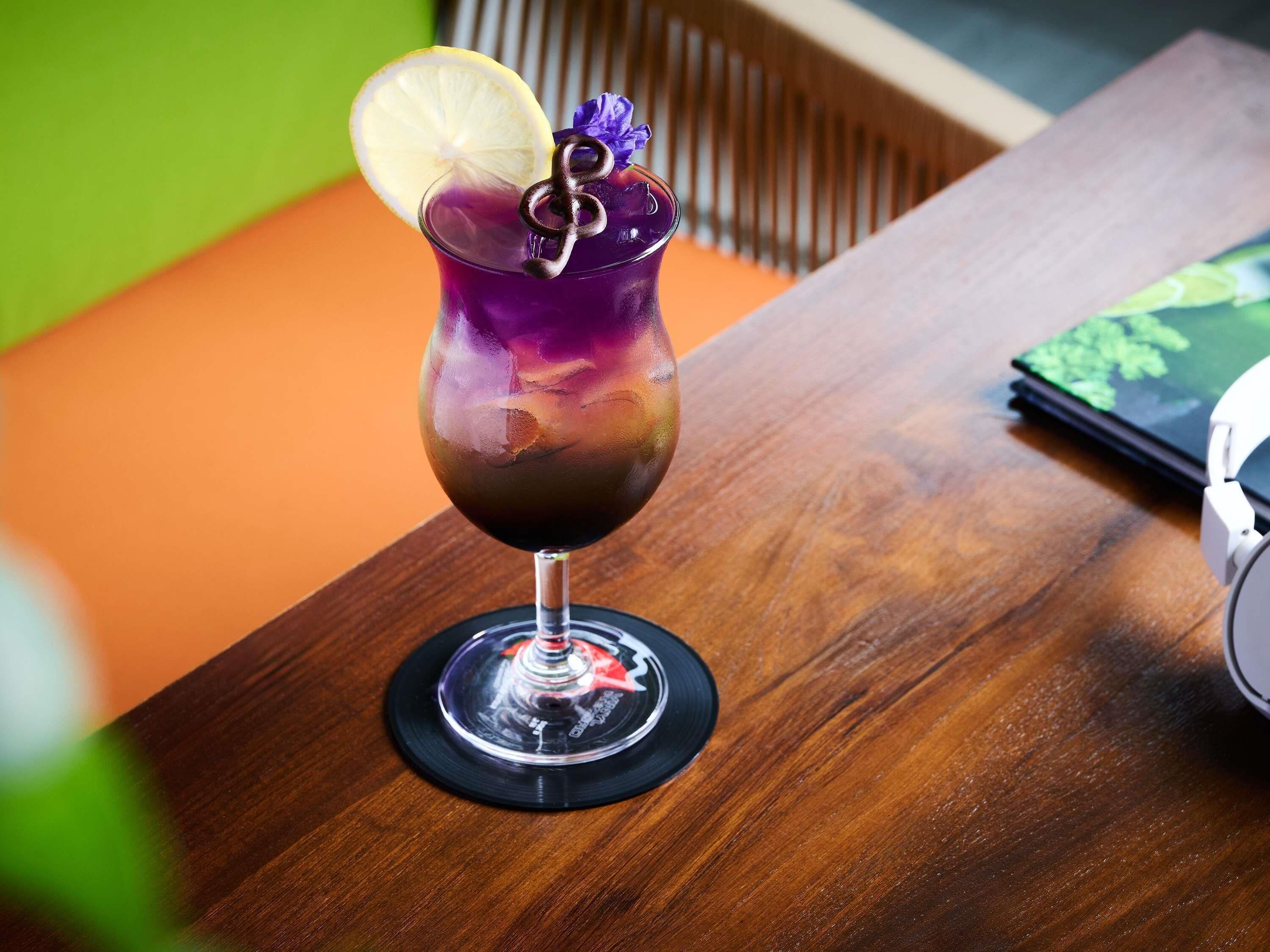 A cocktail in a glass with a lemon slice and a decorative metal piece, on a wooden table with a laptop and headphones nearby.