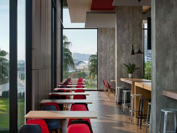 Un comedor moderno con mesas largas y sillas rojas, ventanas amplias con vistas a la ciudad y un pilar de hormigón.