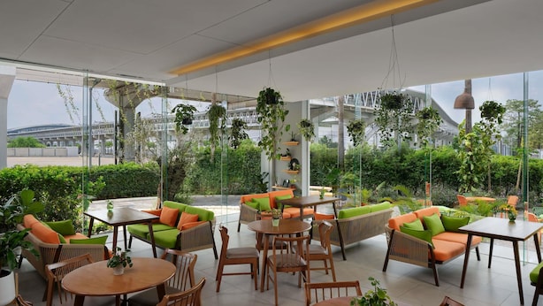 A modern outdoor seating area with tables and chairs, surrounded by greenery and large windows. - ibis Bangkok IMPACT (Pak Kret)