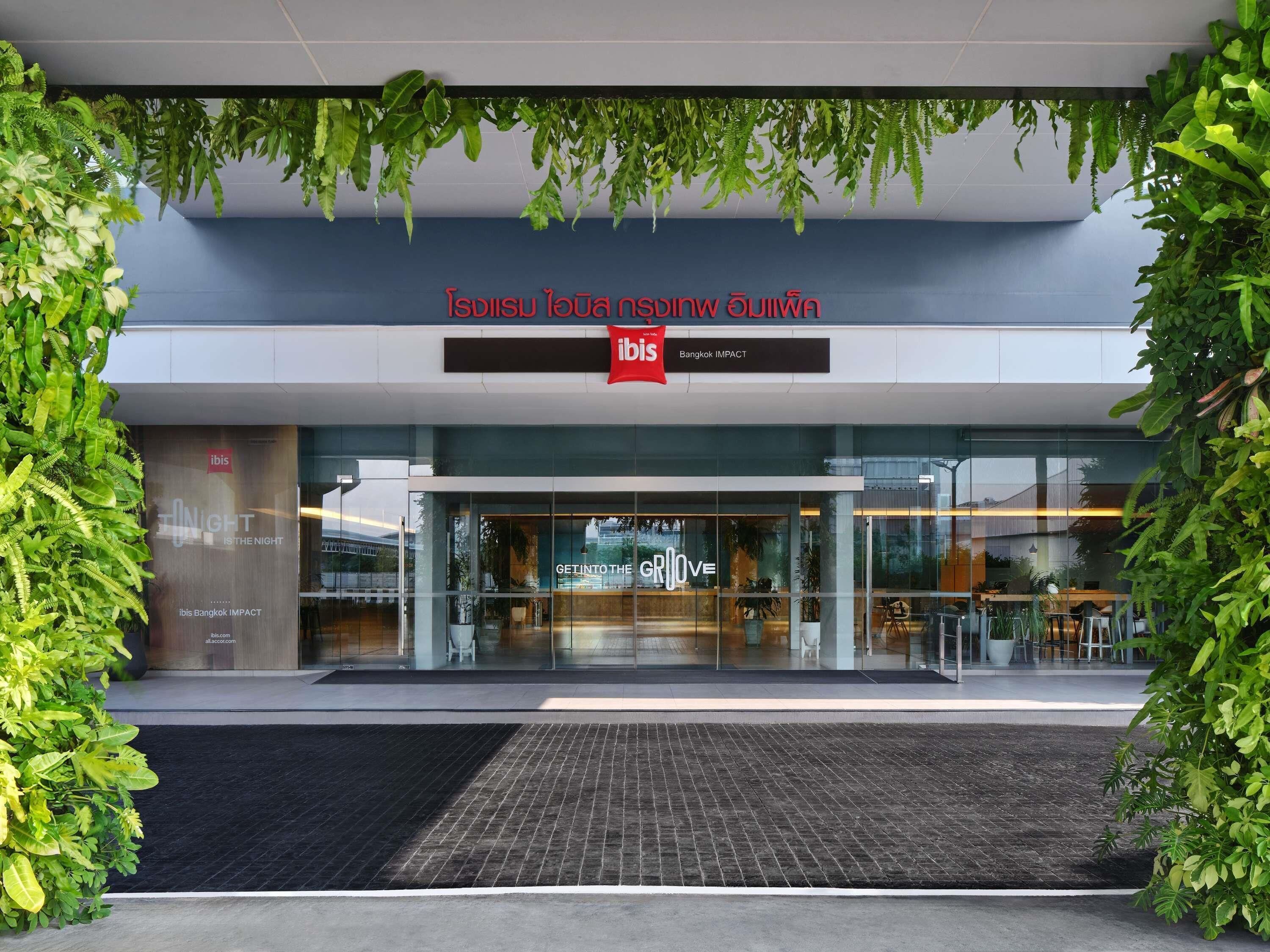 The entrance of ibis Bangkok IMPACT with a modern design, glass facade, and greenery.