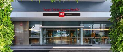 La entrada del ibis Bangkok IMPACT, con un diseño moderno, fachada de vidrio y vegetación.