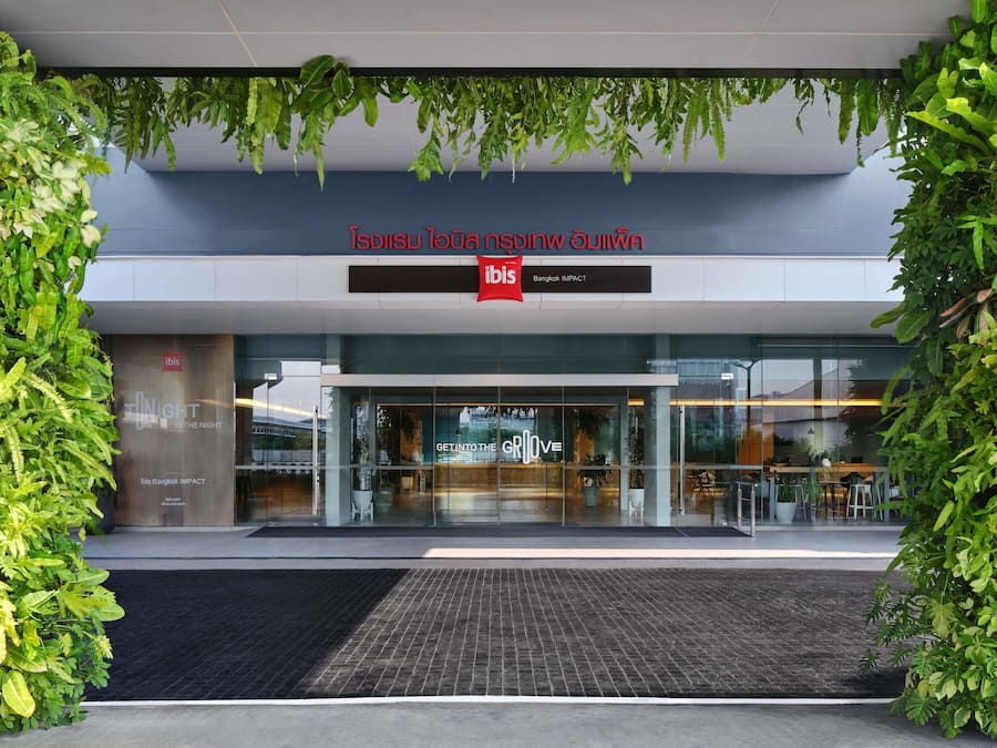The entrance of ibis Bangkok IMPACT with a modern design, glass facade, and greenery.