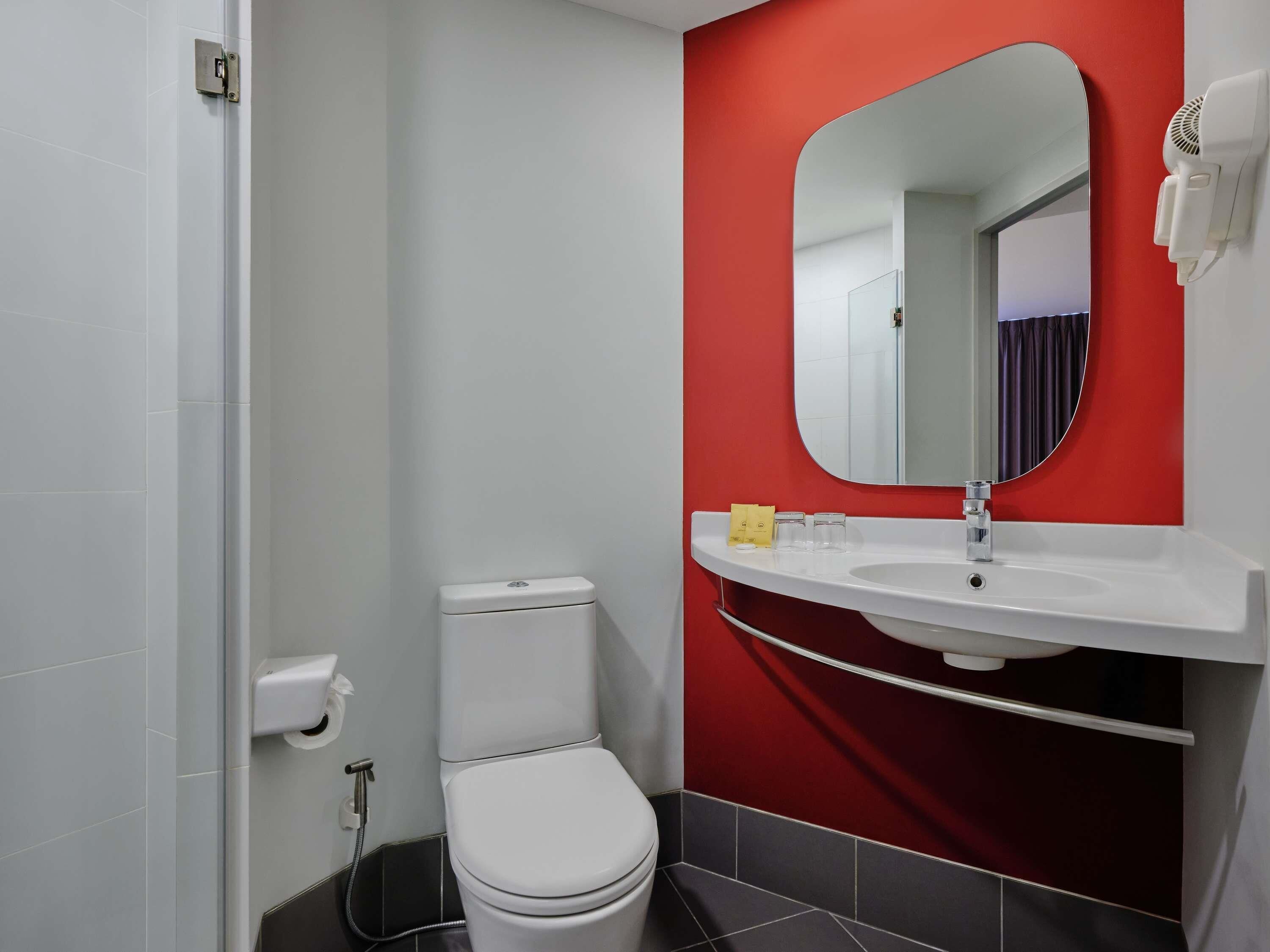 Une salle de bain avec des toilettes blanches, un mur d’accent rouge, un lavabo blanc avec un miroir et un sèche-cheveux fixé au mur.