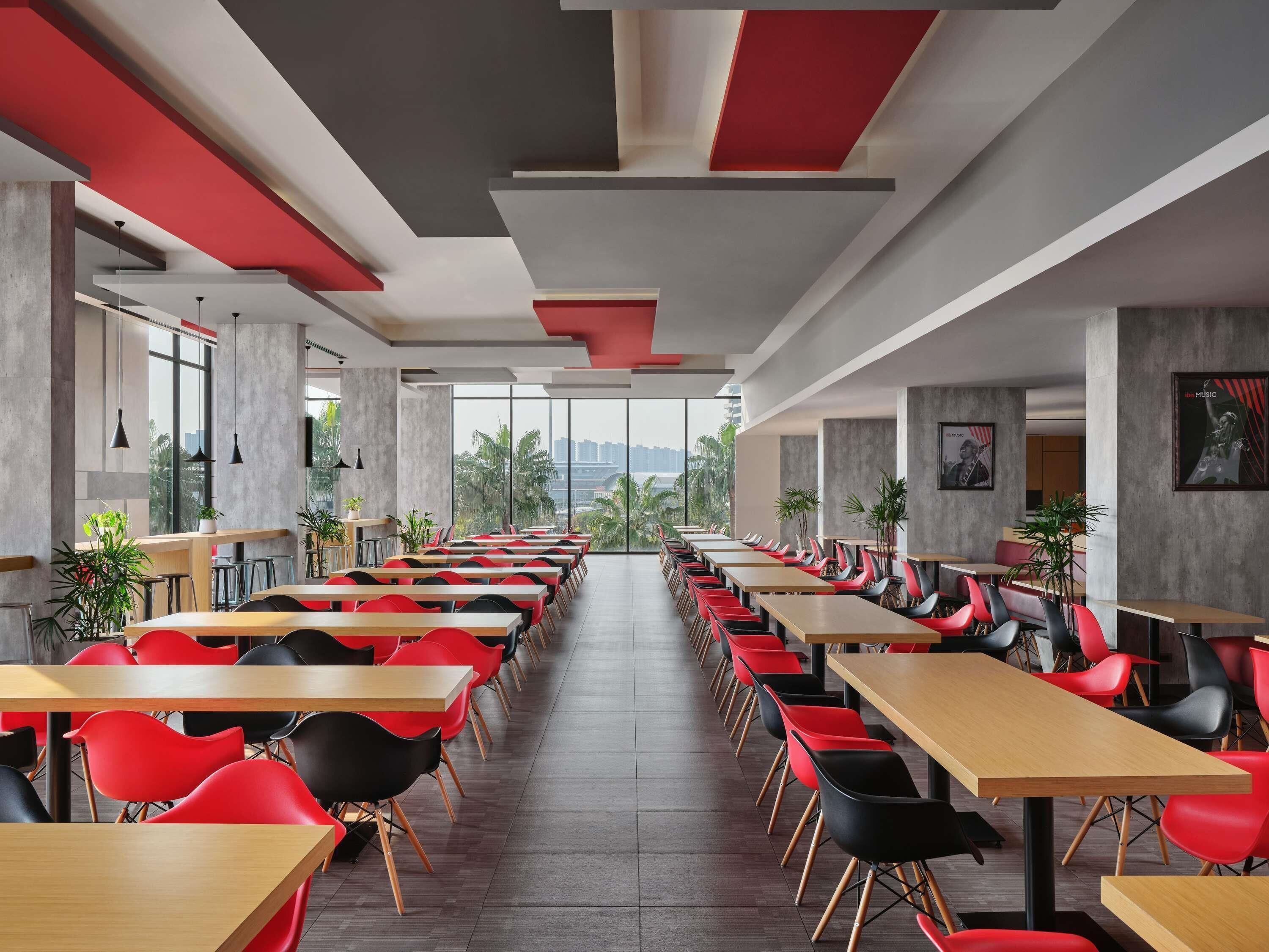 Un comedor moderno con mesas largas y sillas rojas, ventanas amplias y vistas a palmeras.