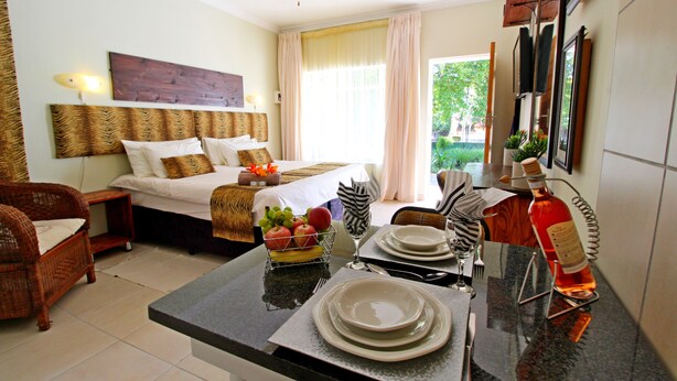 Habitación de hotel con cama, zona de comedor con platos y cubiertos, frutera y botella de vino.