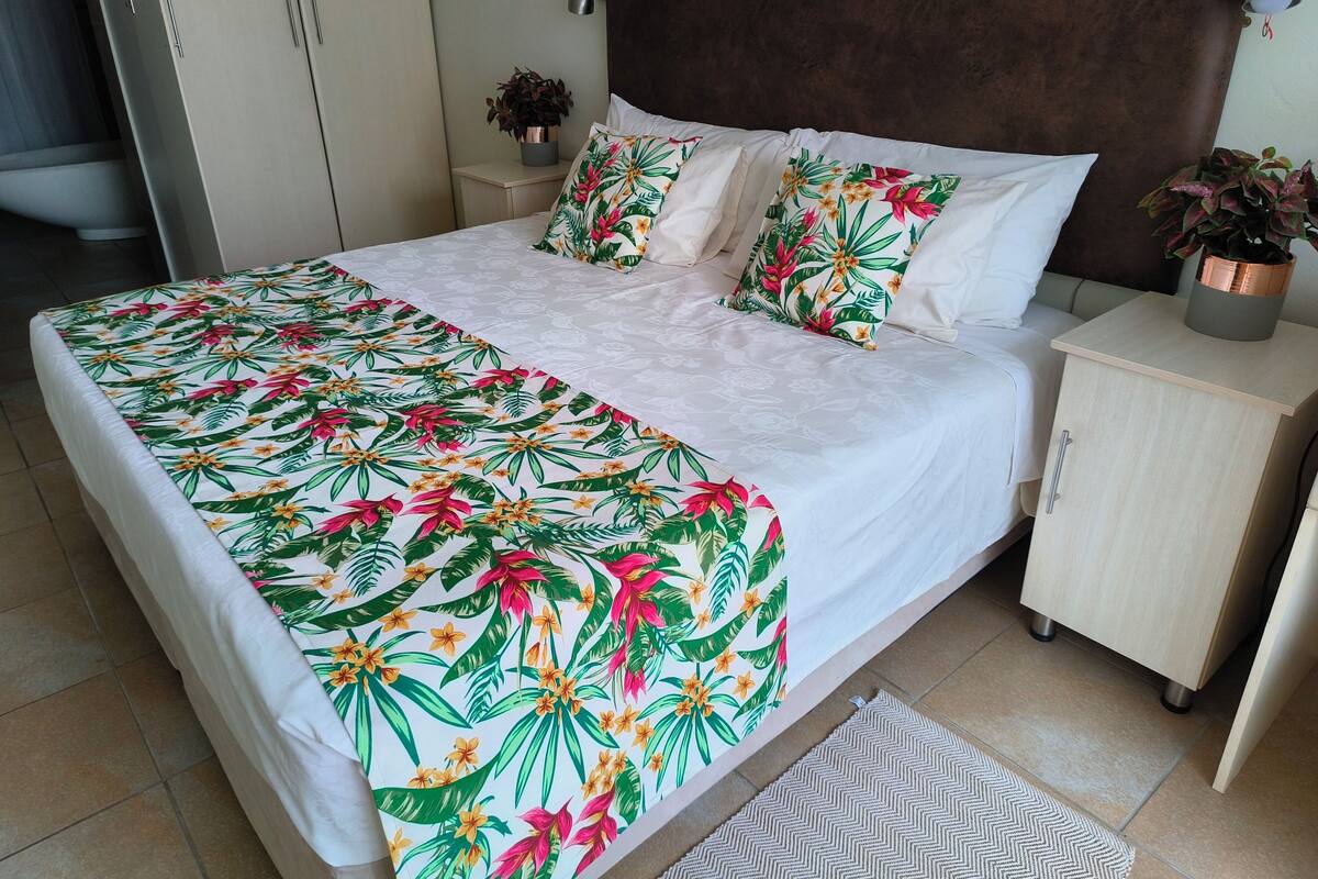 A bed with a floral bedspread, two bedside tables with potted plants, a headboard, and a view of a window with blinds.