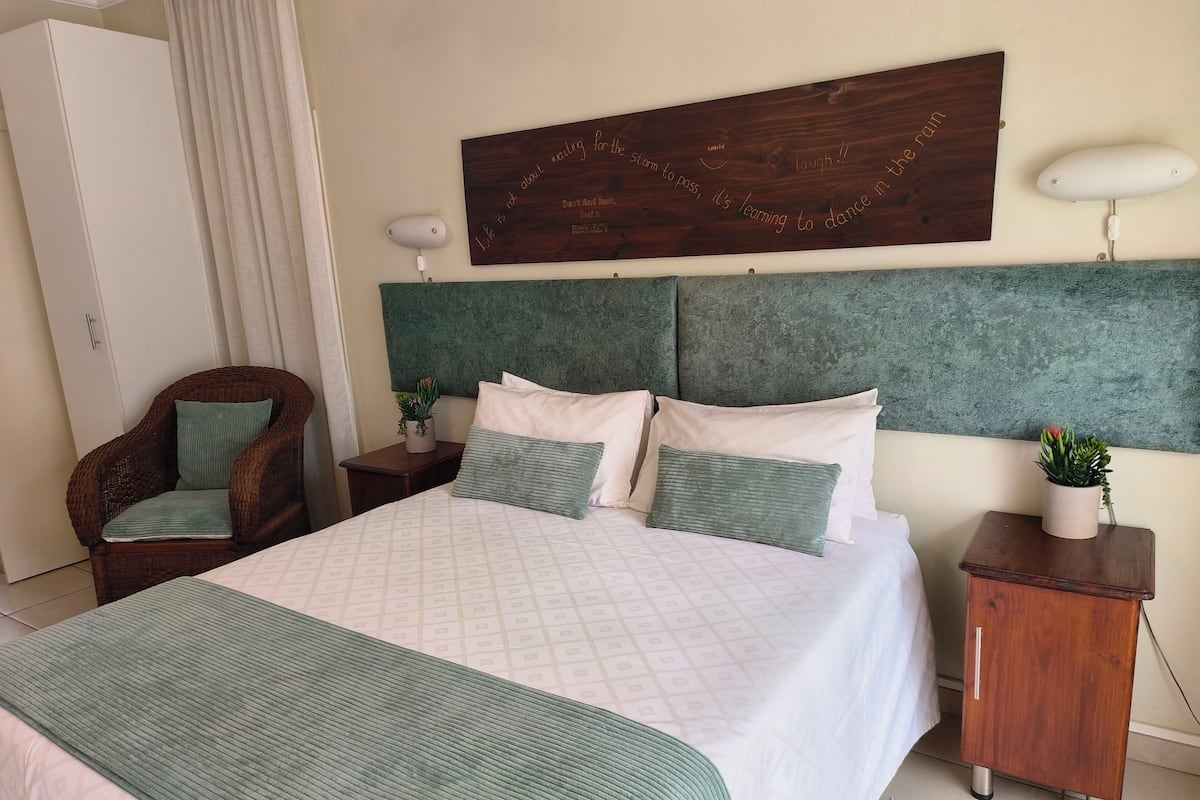 A neatly arranged bedroom with a large bed, a wooden headboard, a chair, a bedside table with a plant, and a wall-mounted lamp.
