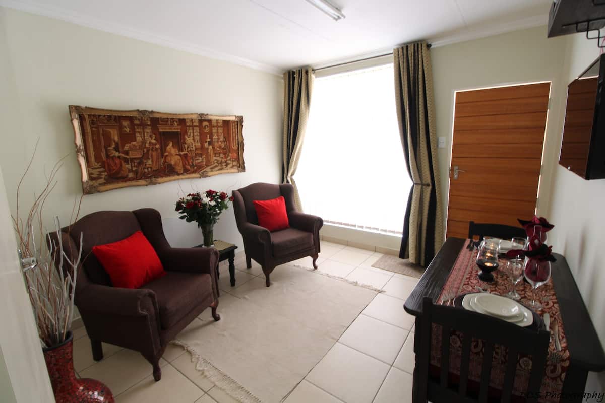 A living room with two brown armchairs, a coffee table with a vase of flowers, a dining table set for two, and a large tapestry on the wall.