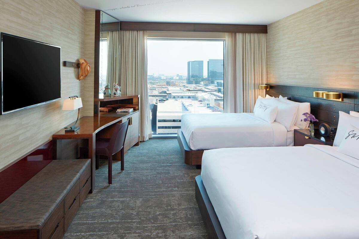 A hotel room with a large TV, a desk, two beds, and a view of the city.