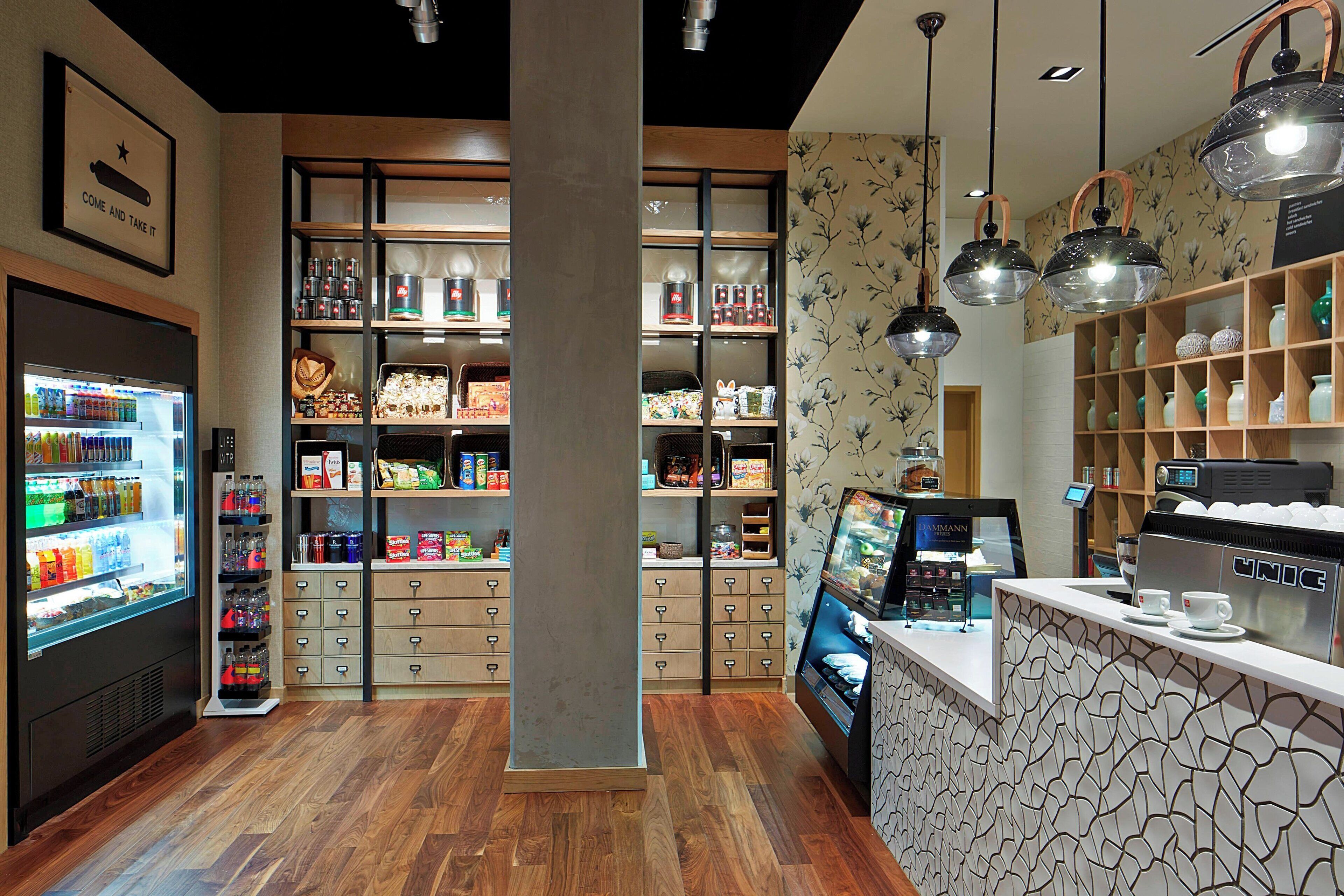 Interior moderno de una cafetería con un expositor refrigerado para bebidas, estantes llenos de snacks, una máquina de café y una pared con diseño.