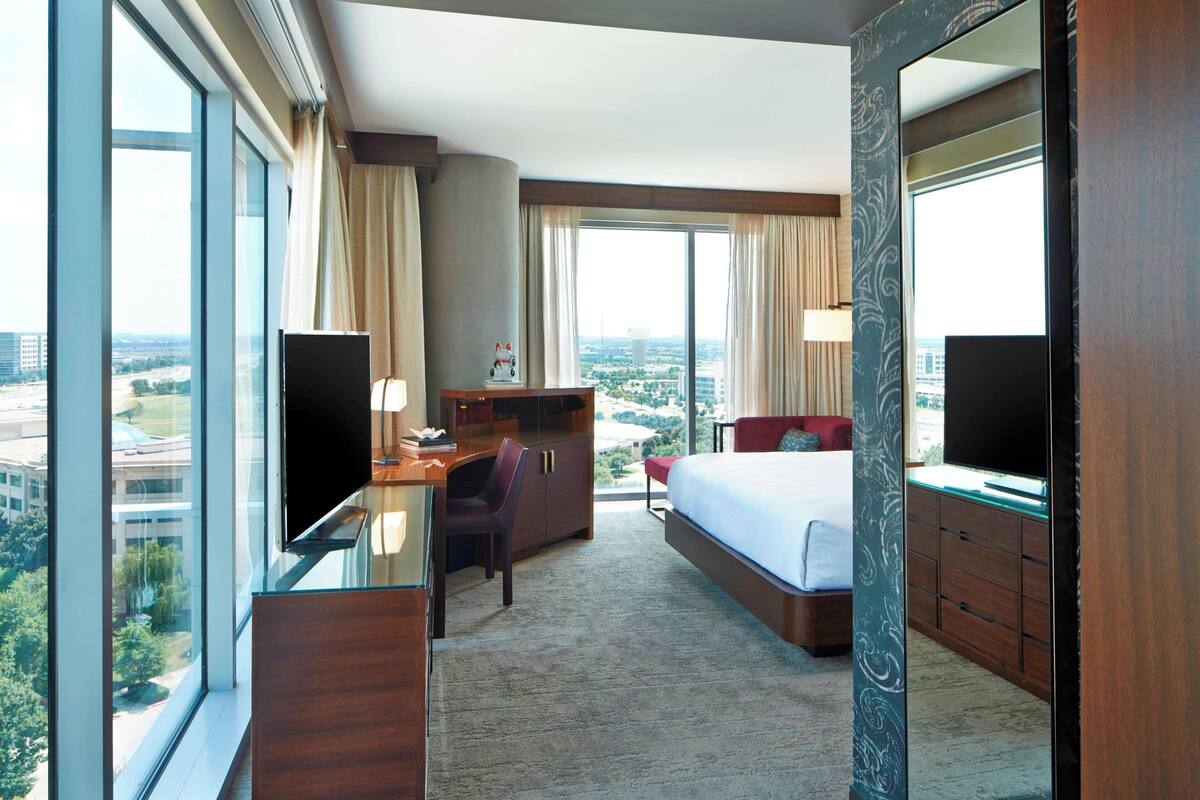A hotel room with a large bed, a desk with a chair, a TV, and a view of the city through large windows.