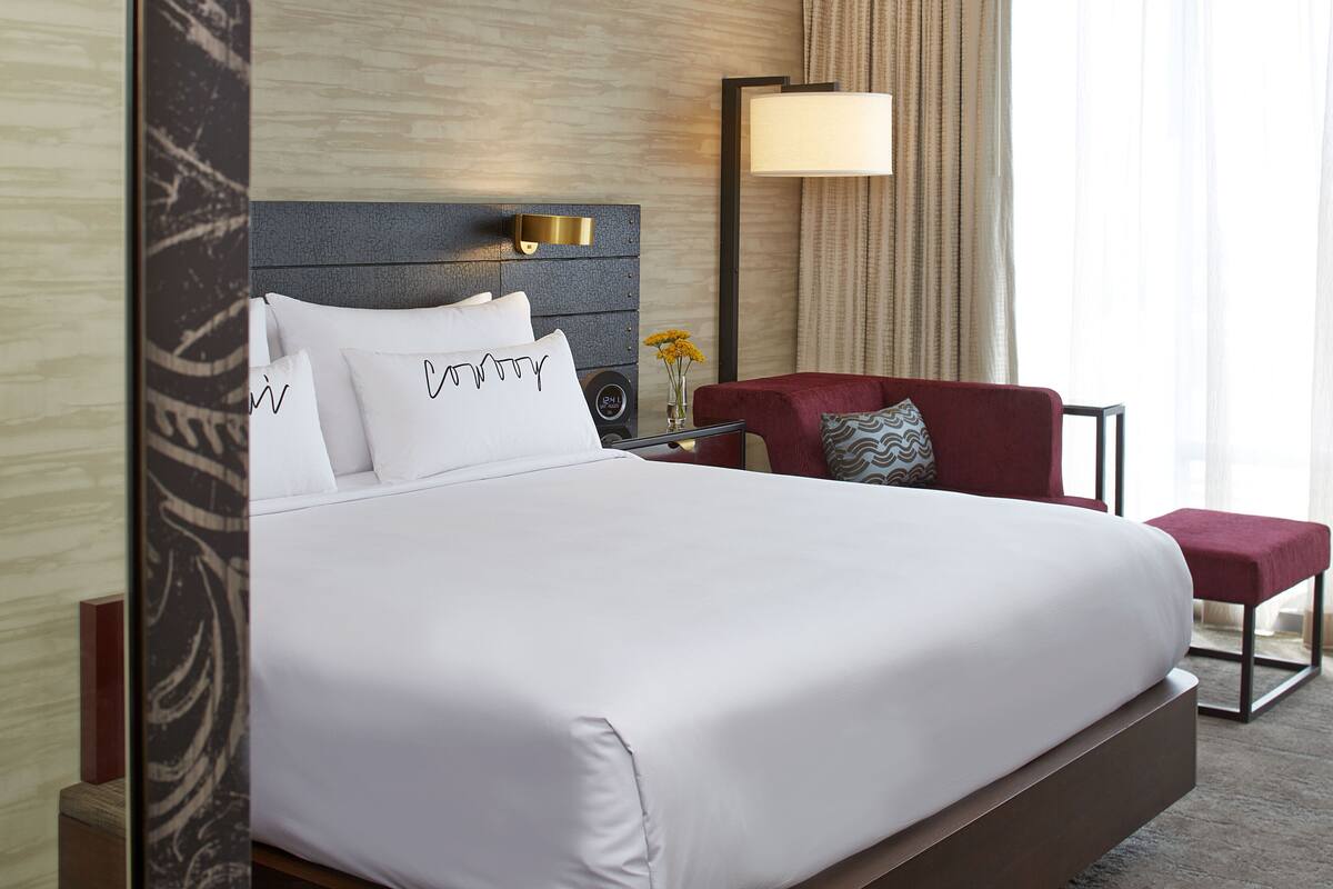 A hotel room with a large bed, a nightstand with a lamp, a red chair, and a small table with a vase of flowers.