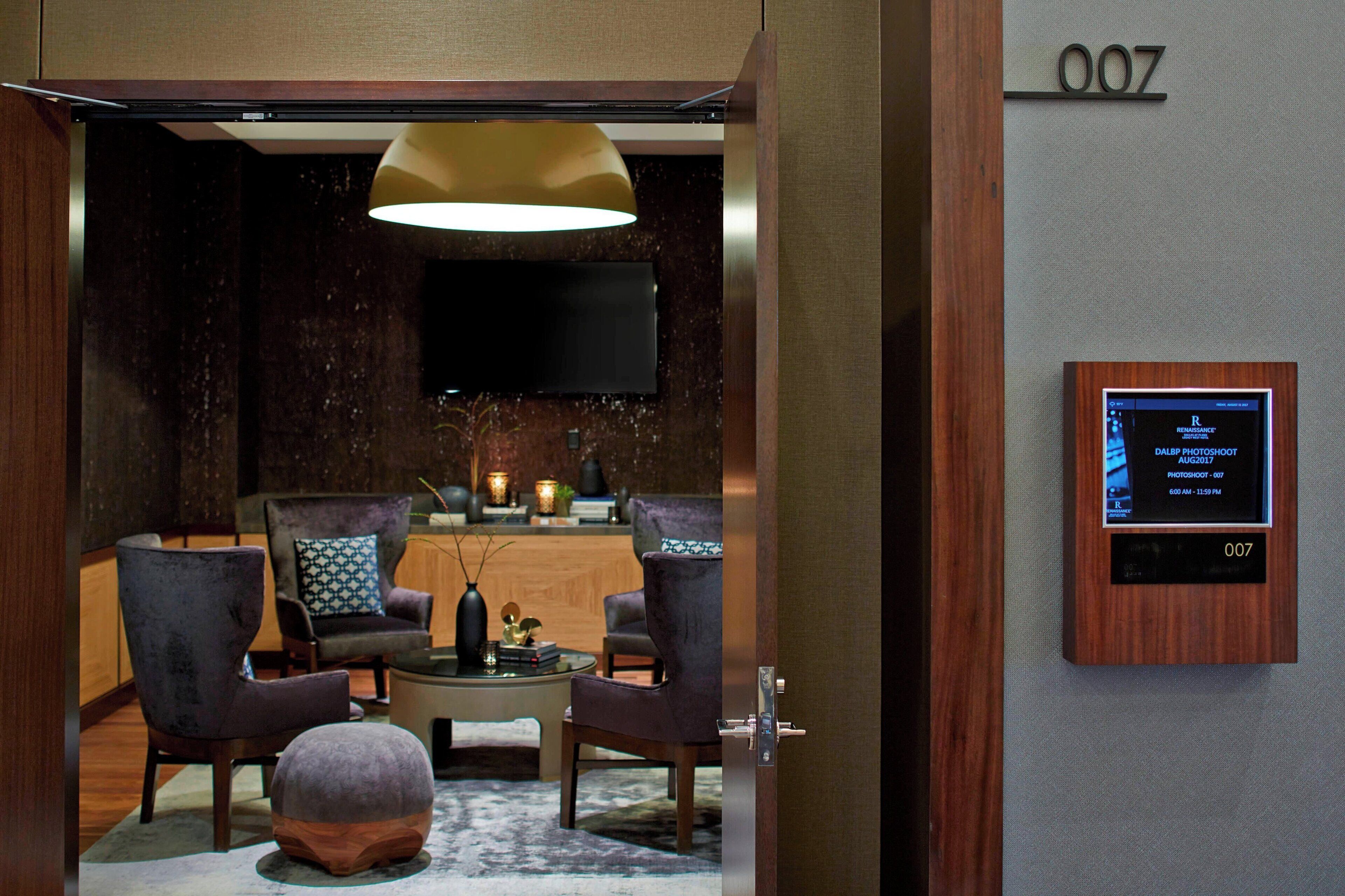 Un moderno vestíbulo de hotel con un mostrador de recepción, dos sillones, una mesa de centro y una lámpara colgante grande. Una pantalla digital muestra detalles de las habitaciones.