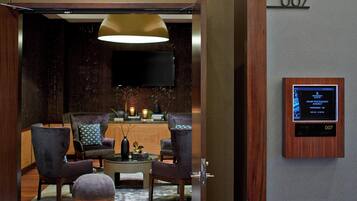 Un moderno vestíbulo de hotel con un mostrador de recepción, dos sillones, una mesa de centro y una lámpara colgante grande. Una pantalla digital muestra detalles de las habitaciones.