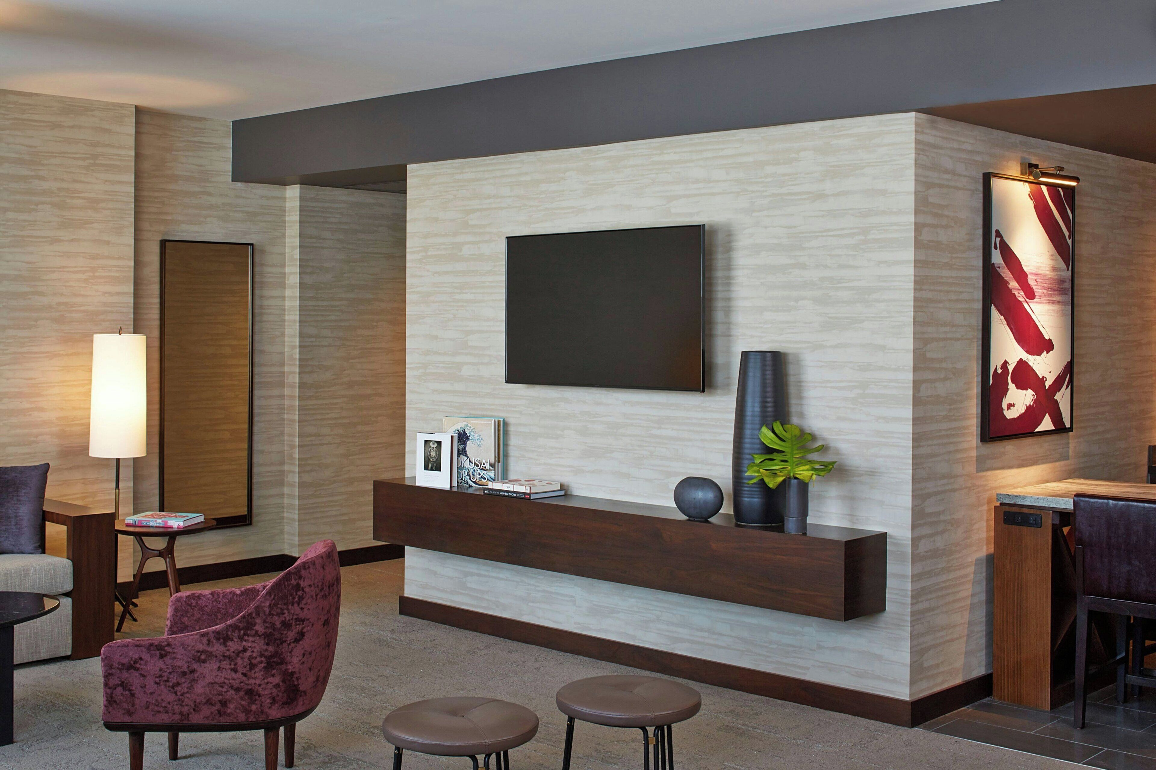 A modern hotel lobby with a flat-screen TV mounted on a wooden panel wall, a dark wooden reception desk, a plush red armchair, two round bar stools, and a large abstract painting.