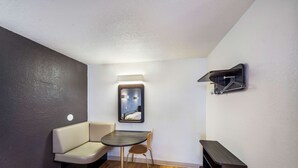 A small, modern room with a dark-colored sofa, a small round table, and a chair. There is a wall-mounted shelf with a black object on it. - Studio 6 Irving, TX - Irving DFW Airport East (Irving)