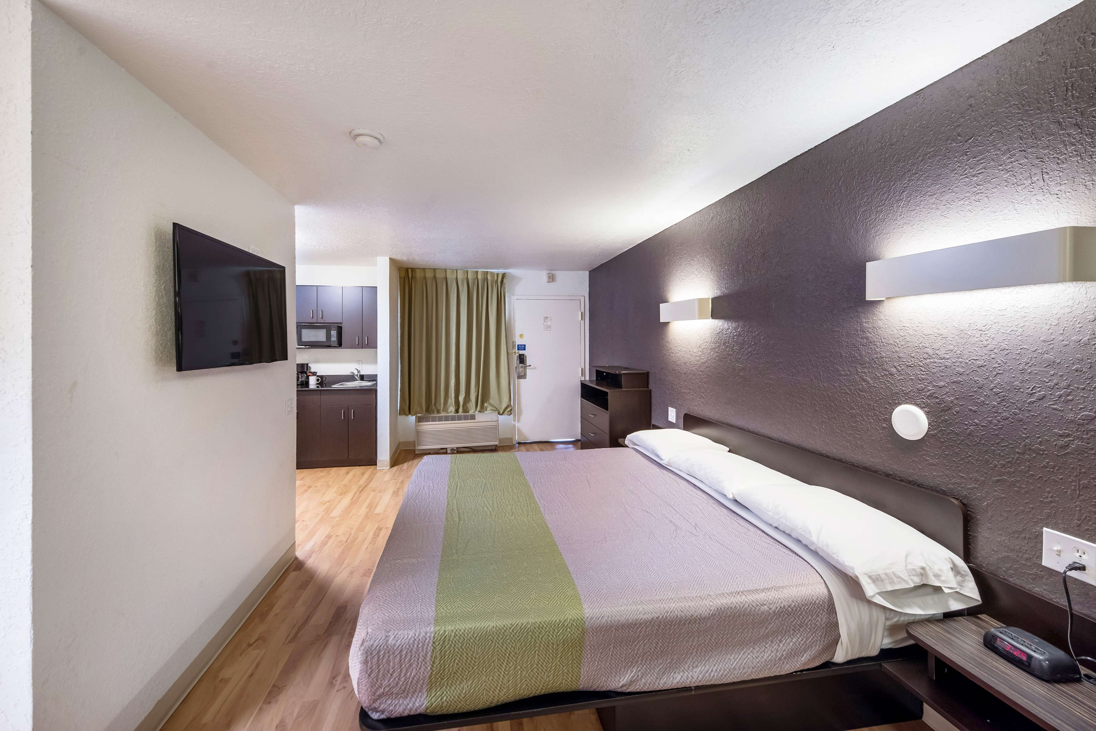 Et hotelværelse med en seng, et natbord, et vægmonteret TV og et lille skrivebord med en lampe.