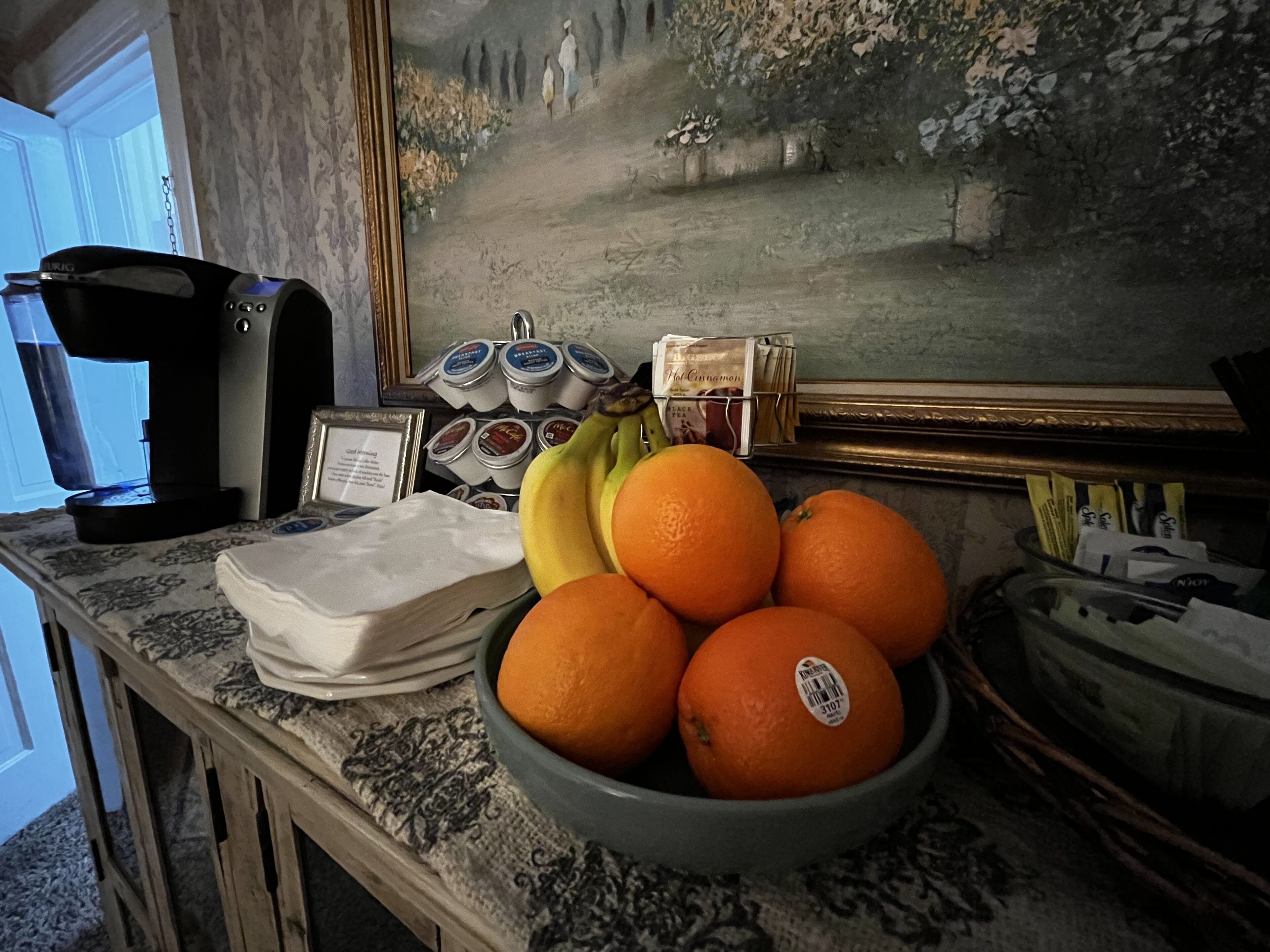A kitchen counter with a coffee maker, a bowl of oranges, bananas, and a framed painting.