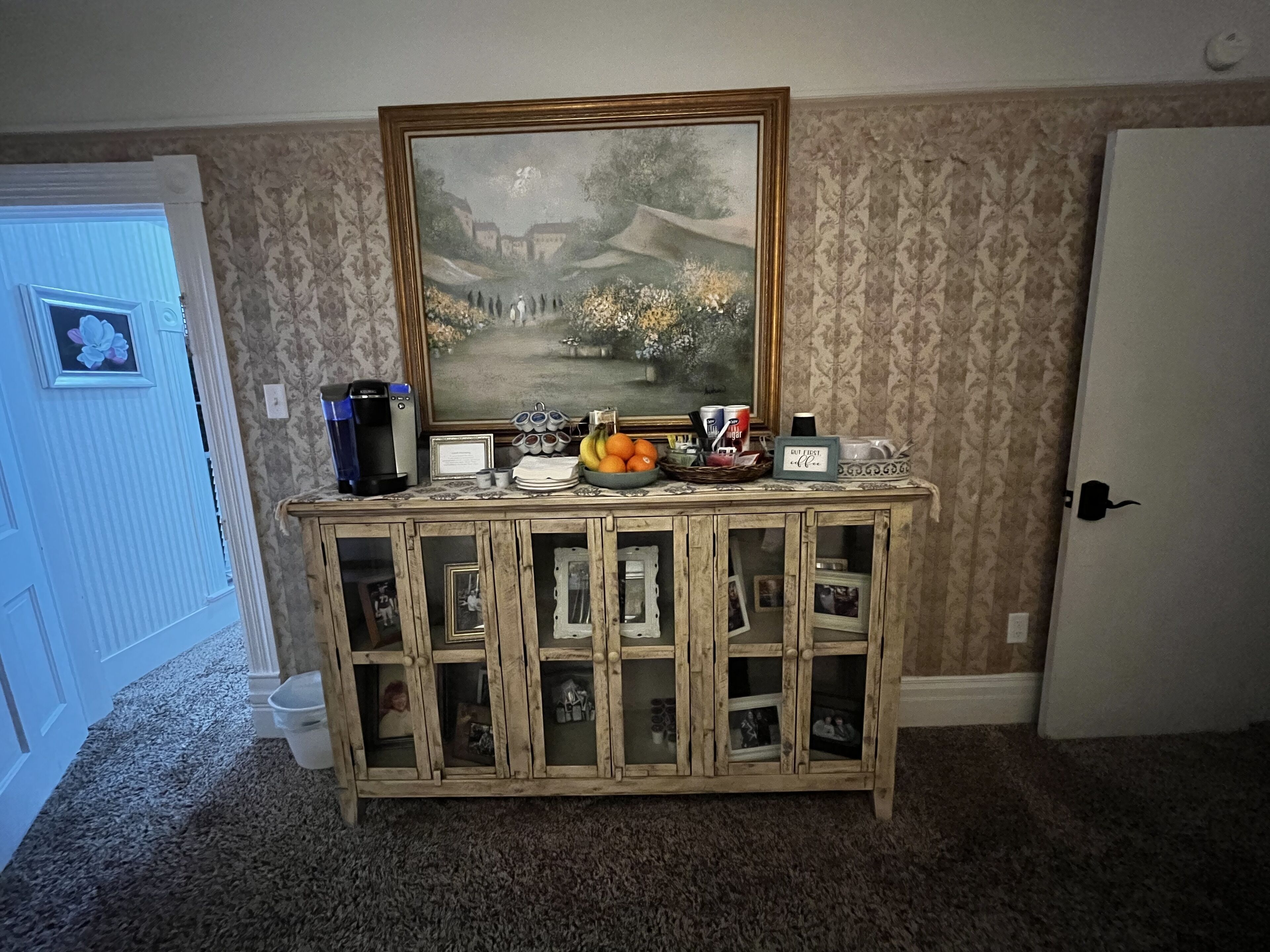 A room with a wooden cabinet featuring glass doors, a coffee maker, a framed painting, and a fruit bowl.