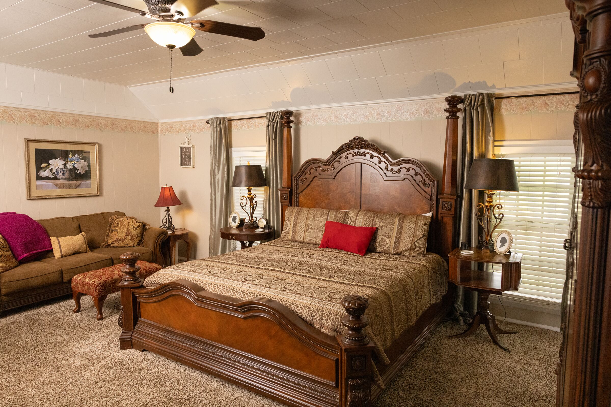 A bedroom with a large wooden bed, a ceiling fan, a nightstand with a lamp, a dresser, and a window with curtains.