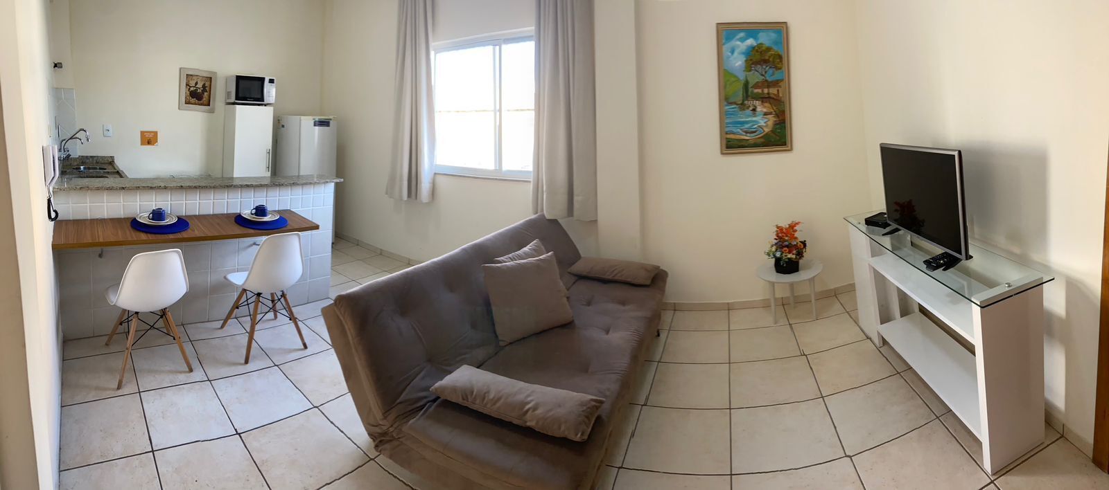 Ein Wohnzimmer mit einer Couch, einem Fernseher auf einem Schrank und einem kleinen Tisch mit einer Vase. Es gibt eine Küchenzeile mit Kühlschrank, Mikrowelle und Spüle.