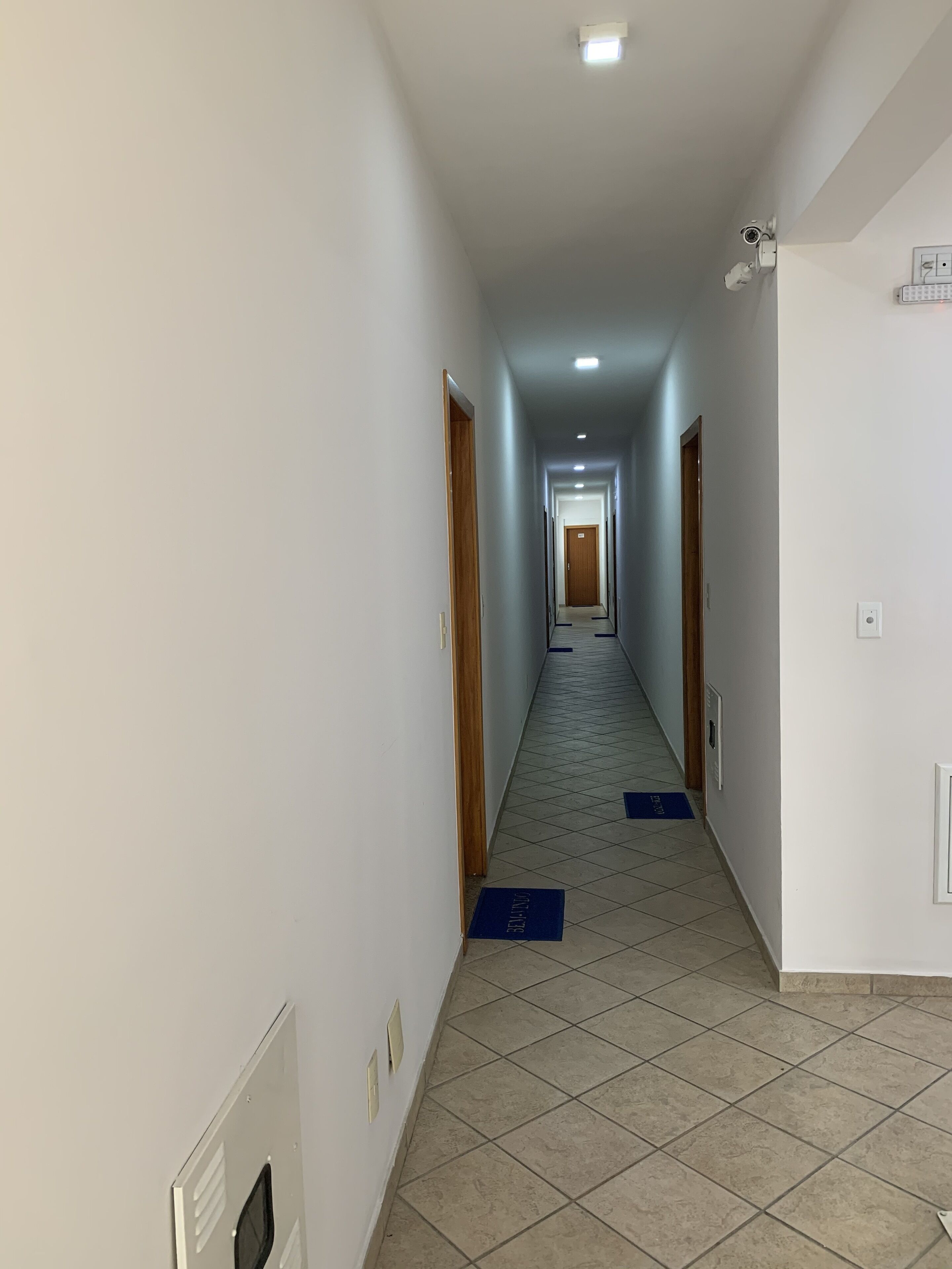 A narrow, tiled hallway with a wooden door at the end, a security camera, and a light fixture on the ceiling.