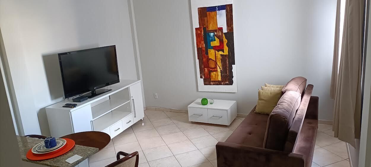 Un salon avec un téléviseur à écran plat posé sur un meuble blanc, un canapé marron, une table d’appoint blanche et un tableau abstrait aux couleurs vives accroché au mur.