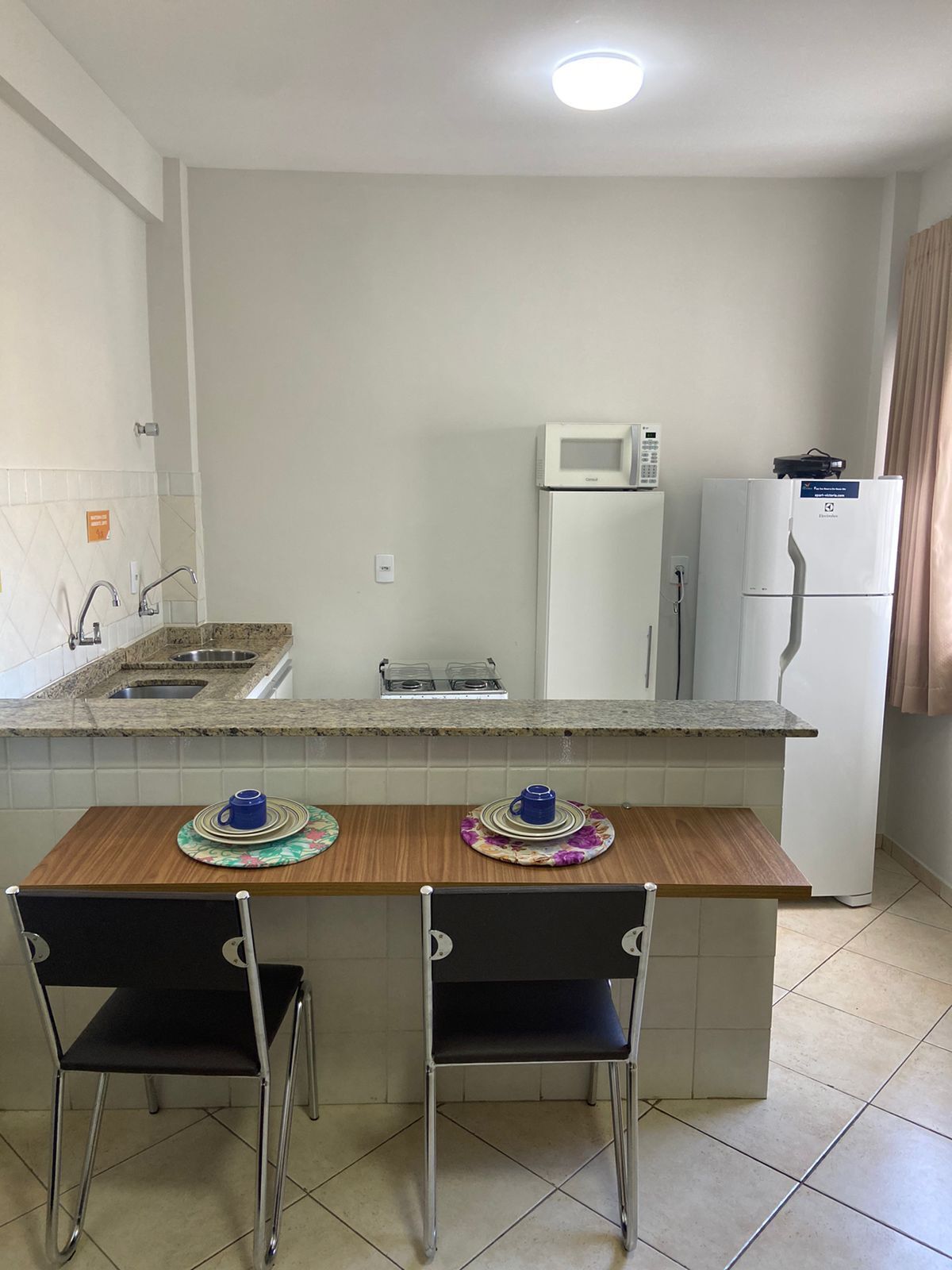 Una cocina compacta con encimera de granito, gabinetes blancos, microondas y refrigerador. Hay dos sillas y una pequeña mesa de comedor.