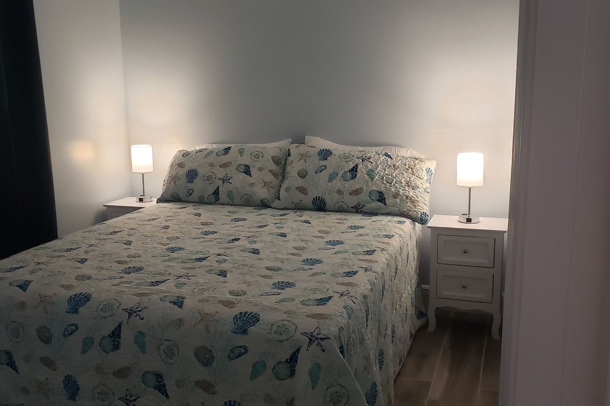 Een net opgemaakt bed met een bloemenbedekking, twee nachtkastjes met lampen, en een donker gordijn aan de linkerkant.