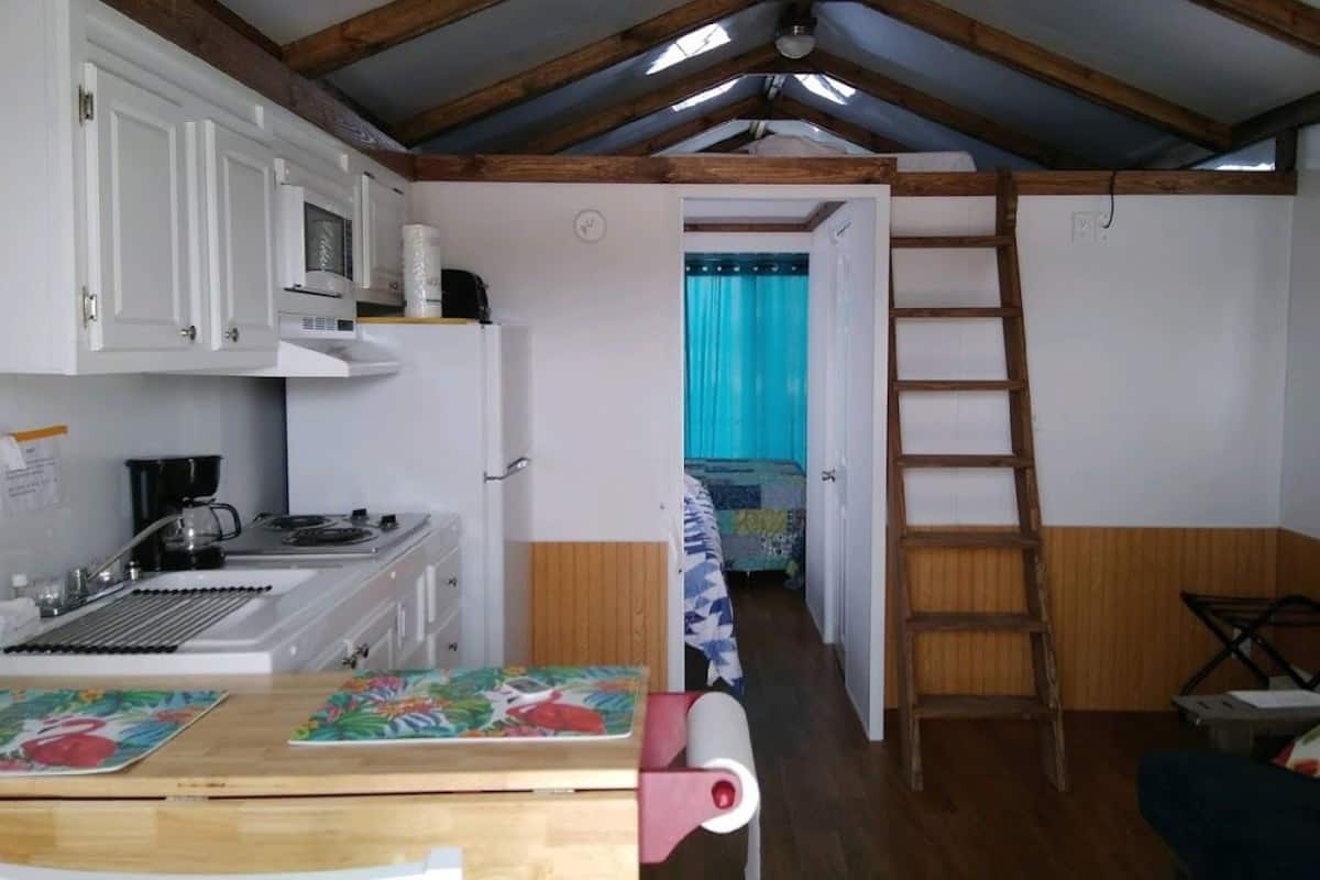 Een kleine keuken met witte kastjes, een magnetron en een spoelbak. Een houten trap staat tegen de muur en er is een bed met een blauw gordijn zichtbaar door een open deur.