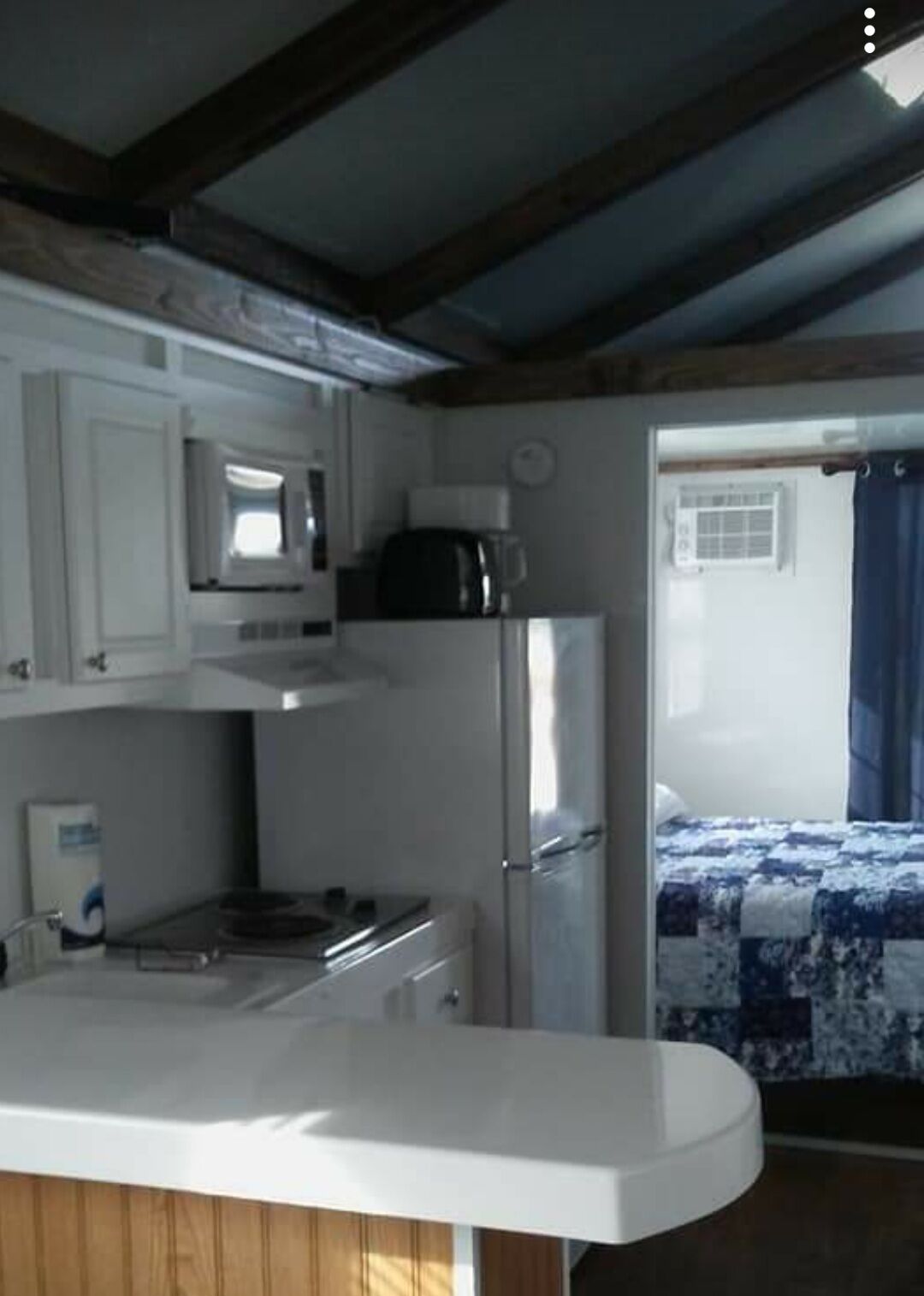 Cocina compacta con armarios blancos, microondas, cafetera y refrigerador. Se ve una cama con colcha de diseño azul y blanco a través de una puerta abierta.