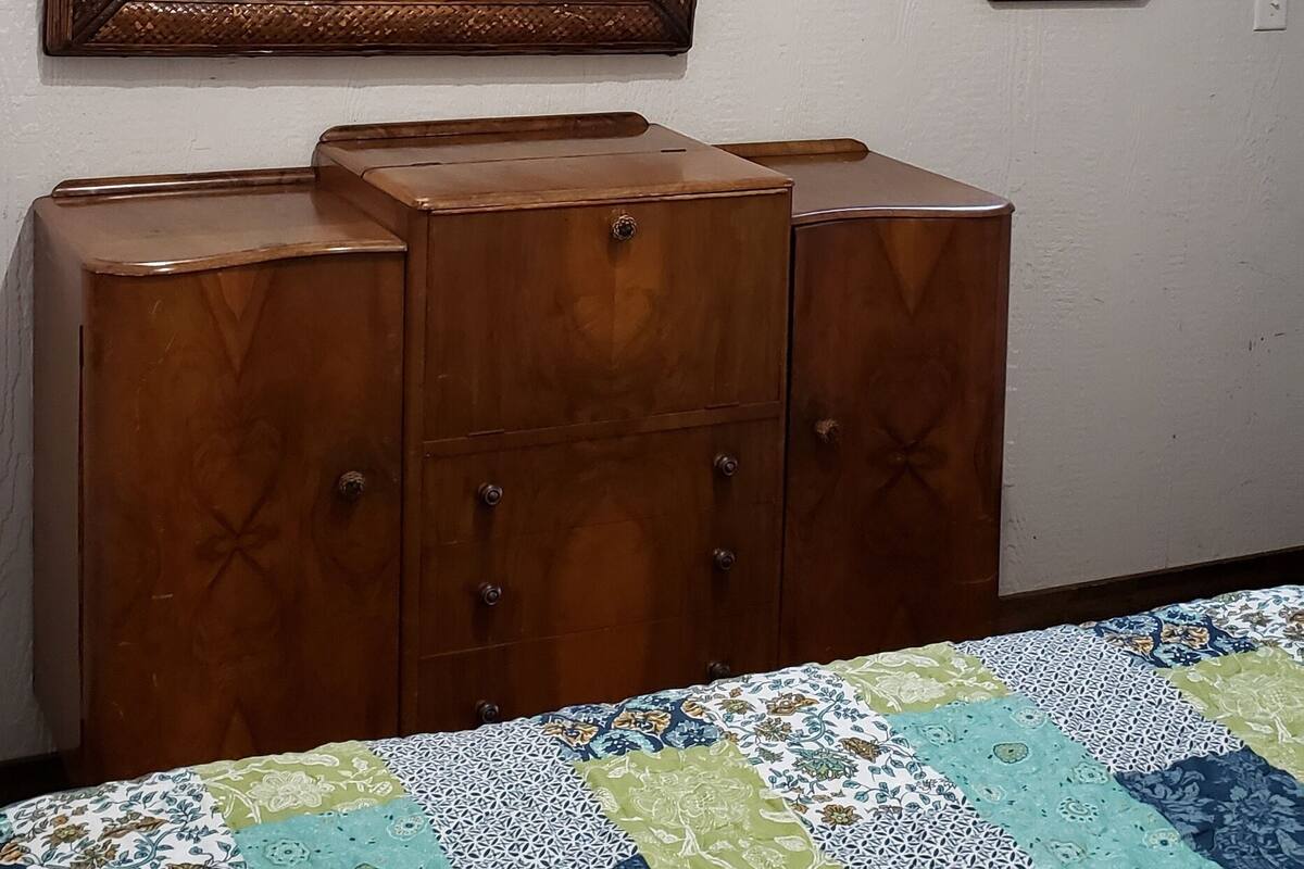 Een slaapkamer met een houten dressoir, een bed met een sprei, een spiegel en een raam met uitzicht op de lucht.