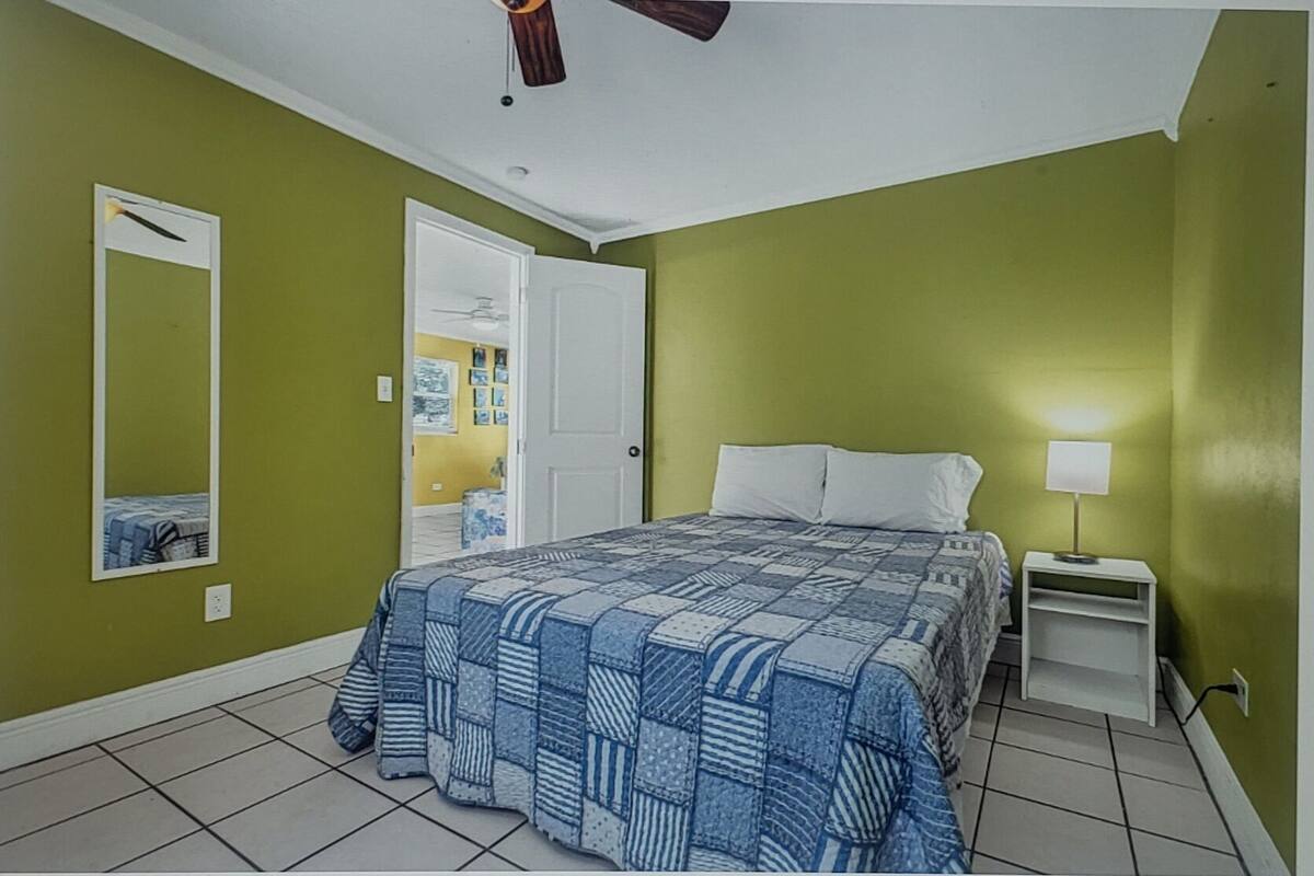 Een slaapkamer met een bed, nachtkastje, lamp, plafondventilator en een deur naar een andere kamer.