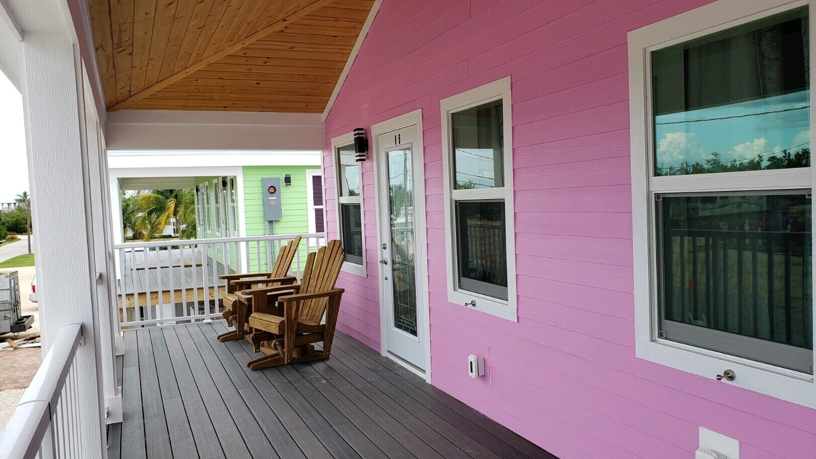 A pink house with a porch, wooden chairs, and a green house in the background.