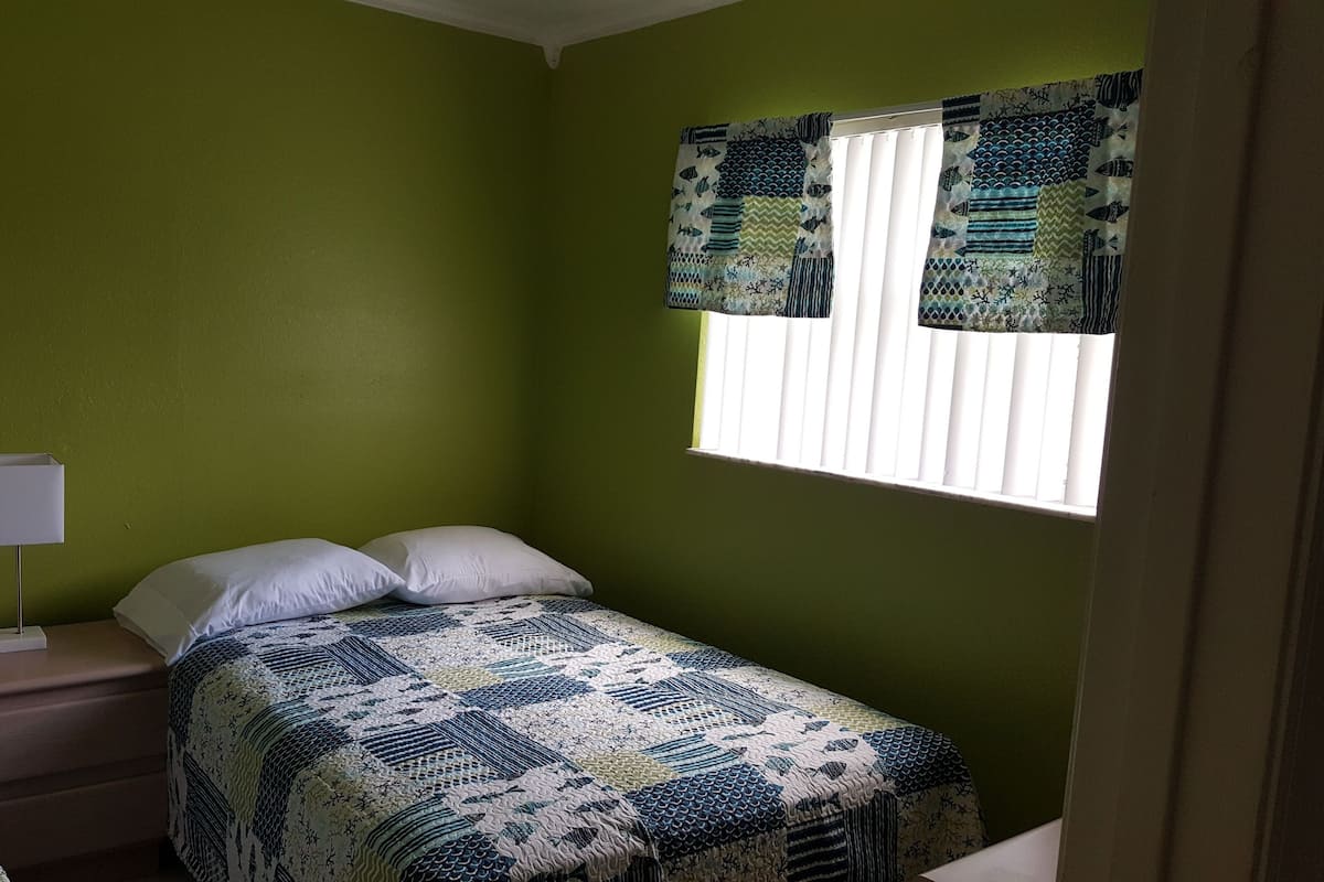 Een kamer met groene wanden, een raam met gordijnen met een patroon, een nachtkastje met een lamp en een bed met een gebedspread met een patroon.