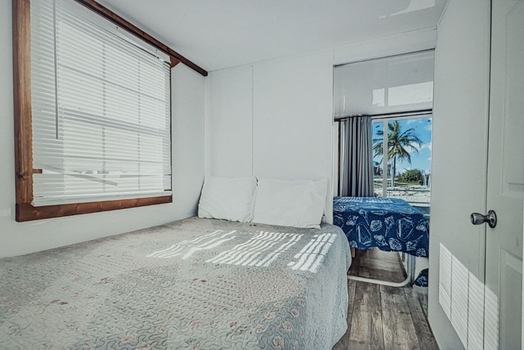 A bedroom with a bed, a window with blinds, a door, and a view of a beach.