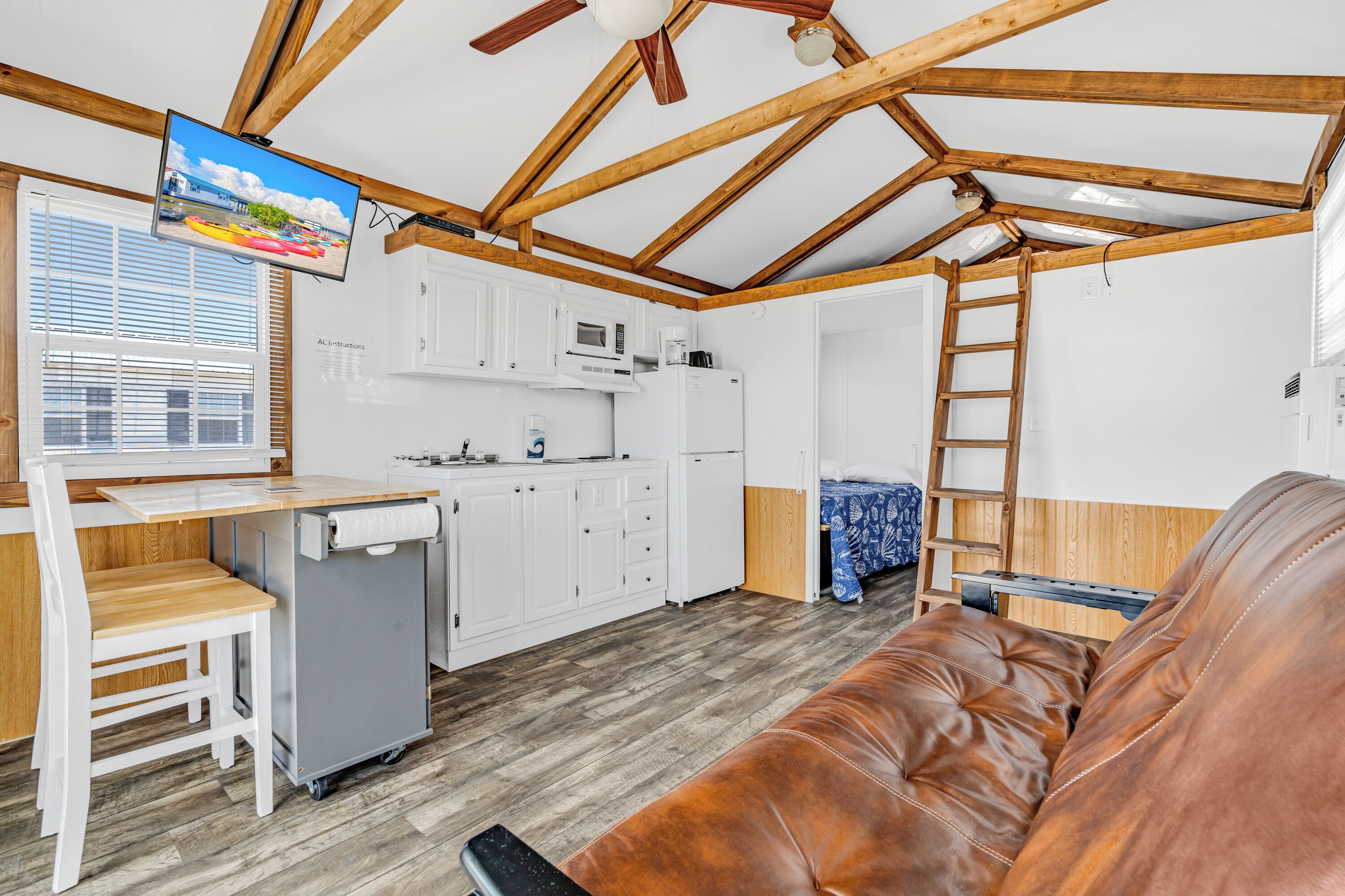 A compact kitchen with white cabinets, a microwave, and a refrigerator. There is a wooden ceiling with exposed beams, a television mounted on the wall, and a brown leather sofa in the living area.