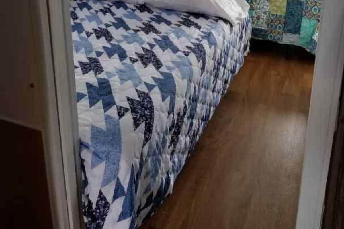 Een slaapkamer met een bed voorzien van een blauw-wit gebeddeken, een houten vloer en een raam met gordijn.