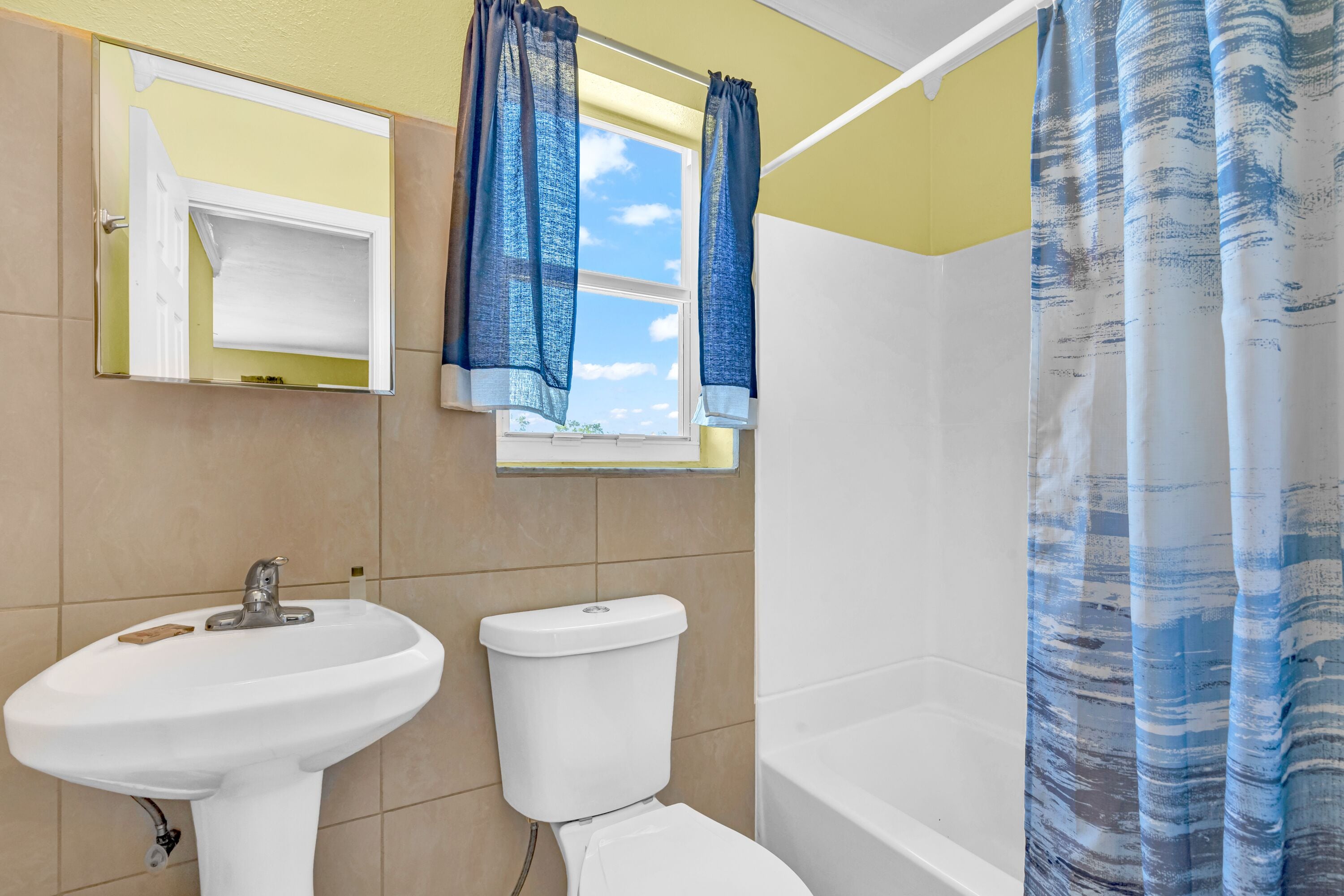 A bathroom with a white sink, toilet, and bathtub. There is a window with blue curtains and a mirror above the sink.